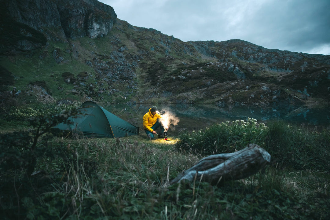 Camper in a yellow jacket lighting a stove beside a green tent at a dark mountain tarn