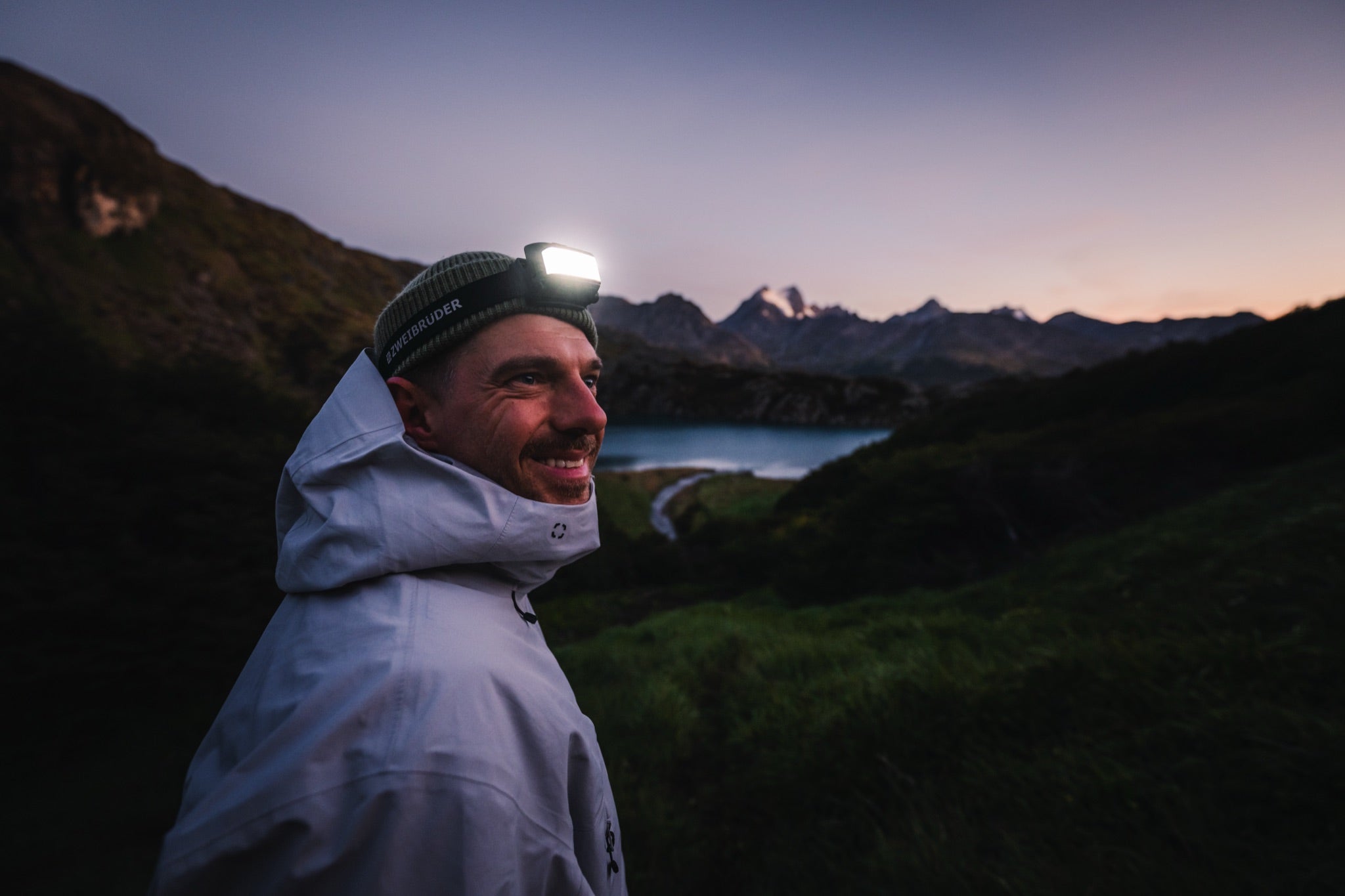 Man wearing a Zweibrüder head torch at dusk by a mountain lake