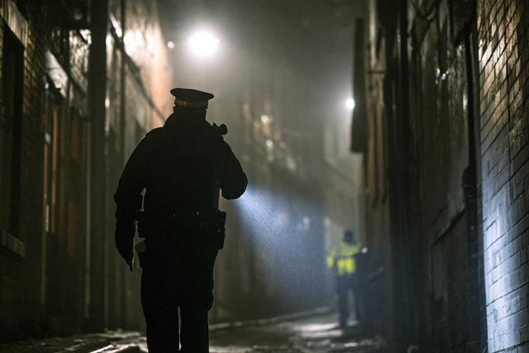 Police officer walking a dark alleyway at night, torch beam lighting the wet ground ahead